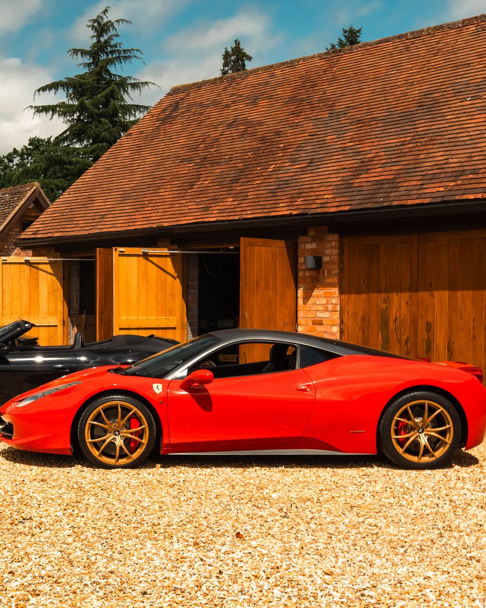 Red Ferrari 458 with gold wheels beside two Porsches outside an oak-doored barn